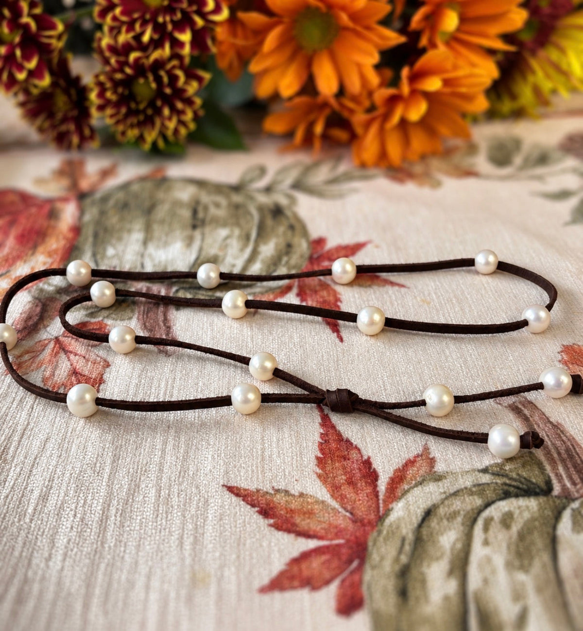 White pearls spaced along a brown leather necklace on autumn leaf-patterned fabric, with orange and burgundy flowers in the background.