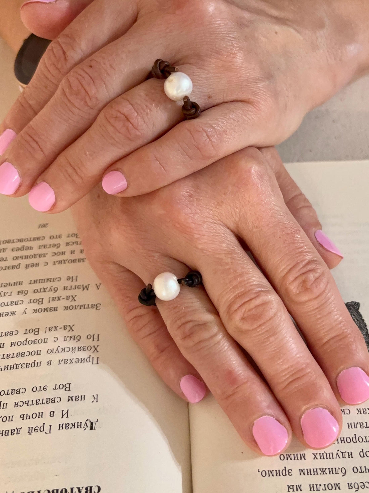 Close-up of hands with pink nail polish wearing pearl rings on a book page.
