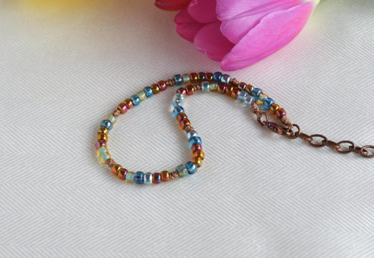Multicolored beaded anklet on a white surface with a pink flower in the background