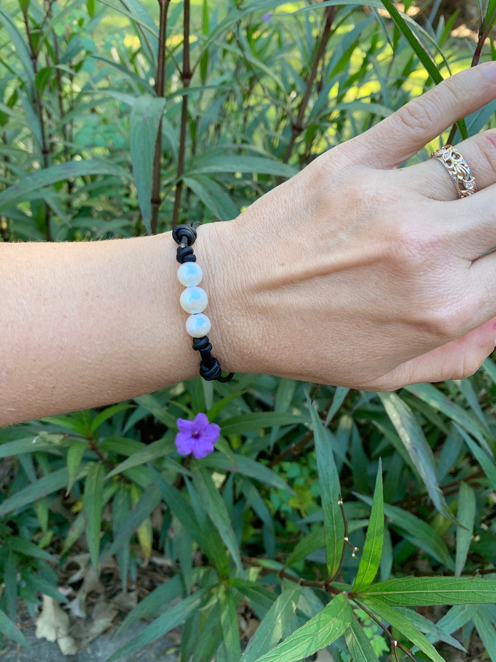 Hand wearing a bracelet with three white pearls against a green leafy background