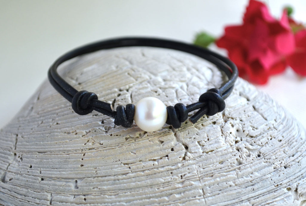 Black cord bracelet with a pearl on a textured surface with red flowers in the background