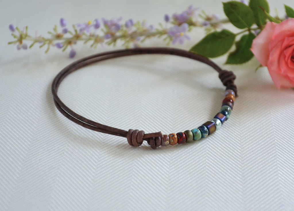 Anklet with colorful beads on a brown cord, placed on a white surface with flowers in the background.