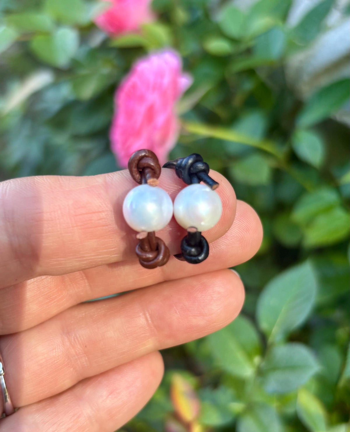 Hand holding a pair of rings with white pearls against a blurred green and pink background