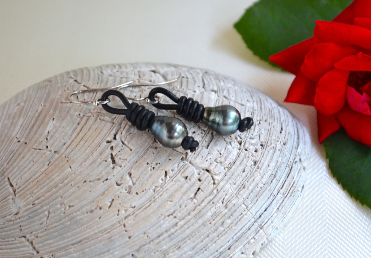 Pair of black Tahitian pearls on black leather cord earrings on a textured surface with a red flower in the background.