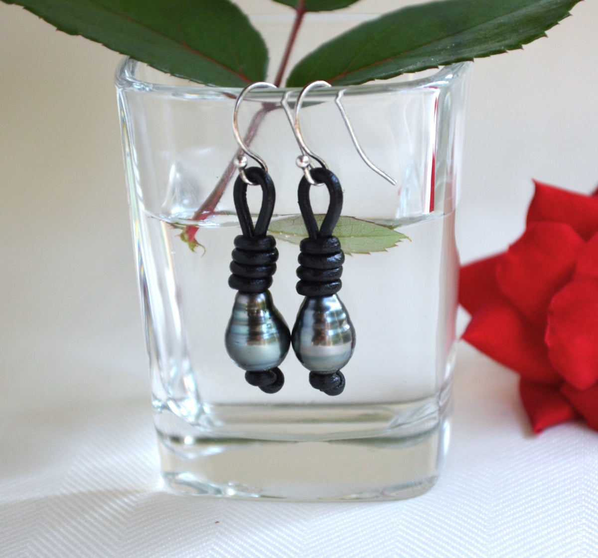 Pair of black Tahitian pearls sterling silver earrings hanging in a clear glass with a red flower and green leaves in the background.