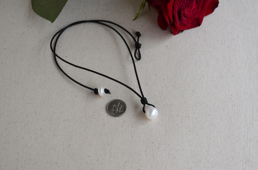Black cord necklace with a pearl pendant on a textured surface with a red rose in the background
