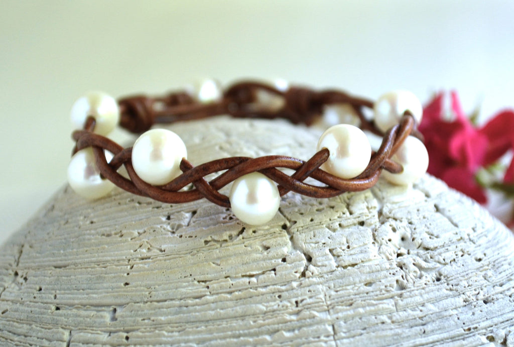Brown braided bracelet with white pearls on a textured surface with flowers in the background