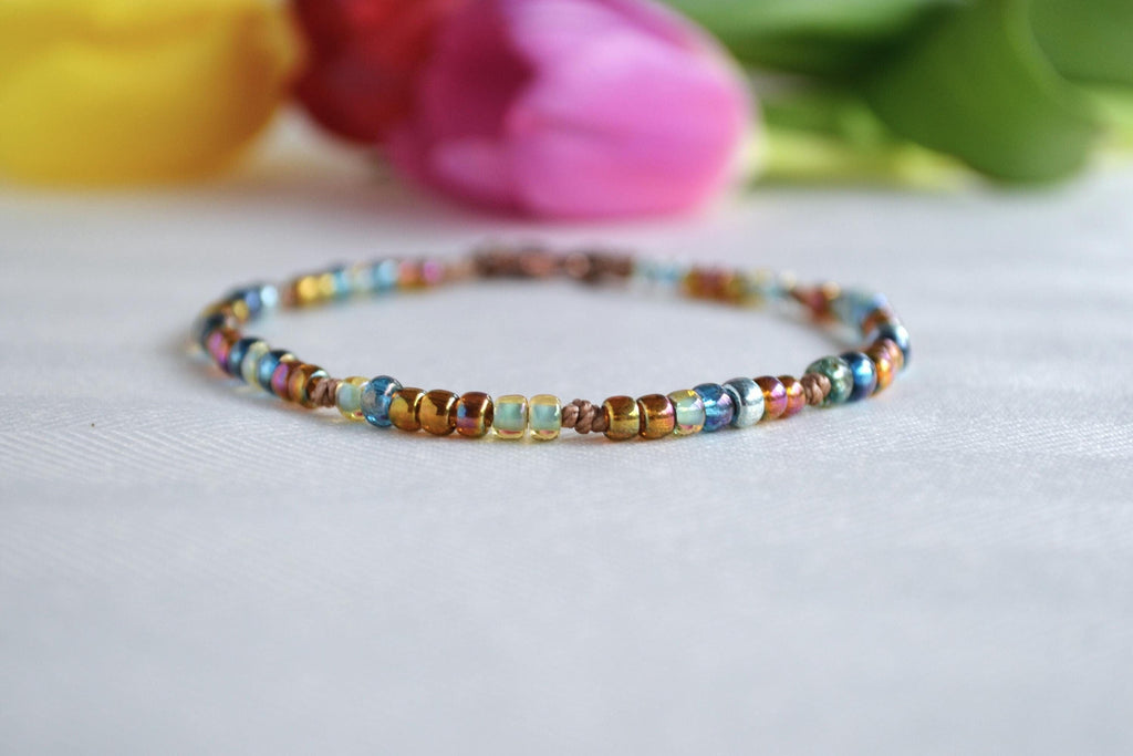 Multicolored beaded ankle bracelet on a light surface with blurred colorful flowers in the background
