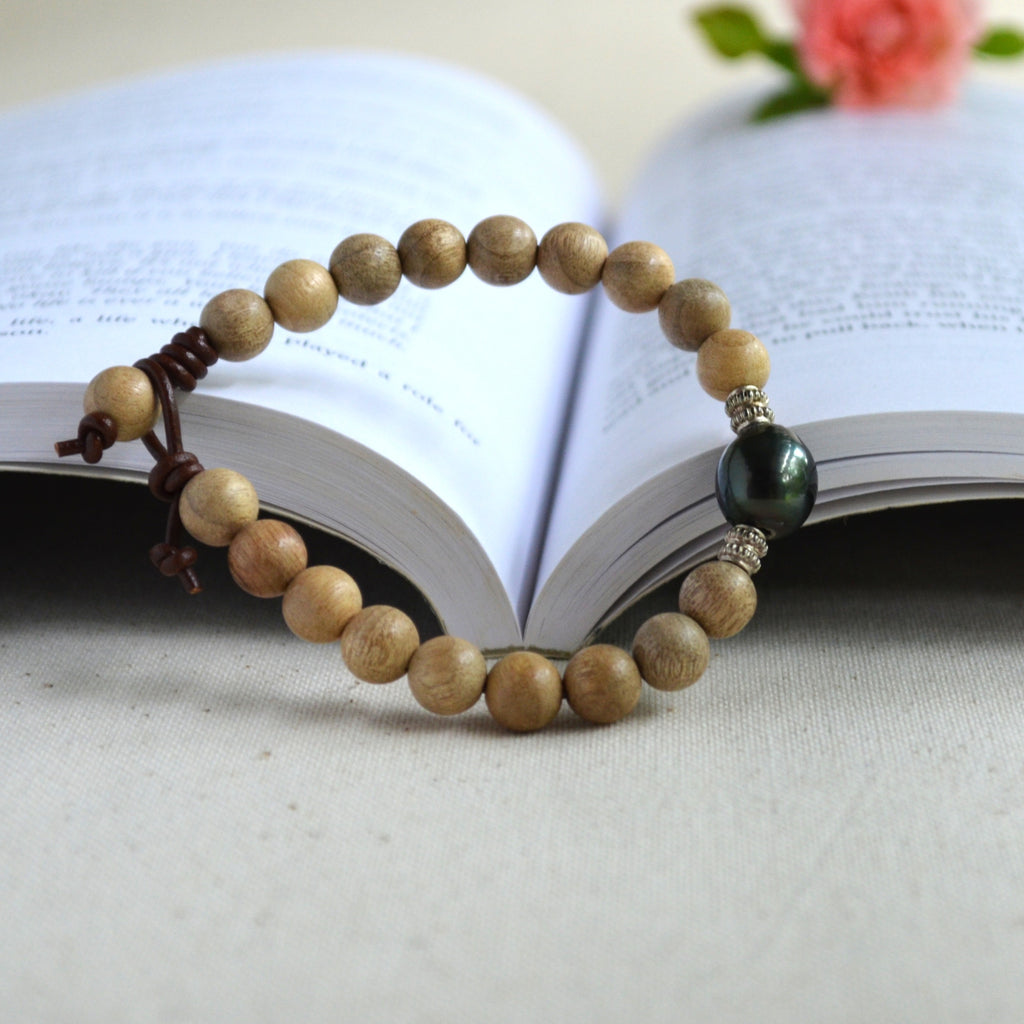 A beaded bracelet made of light brown sandalwood beads with a dark black Tahitian pearl and sterling silver spacer, resting on an open book.