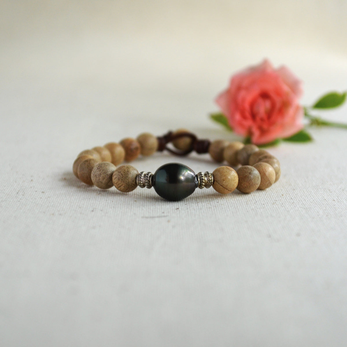 A beaded bracelet with light brown sandalwood beads, a dark black Tahitian pearl, and small sterling silver spacer beads, on brown Greek leather, displayed on a textured white fabric with a soft pink rose in the background.