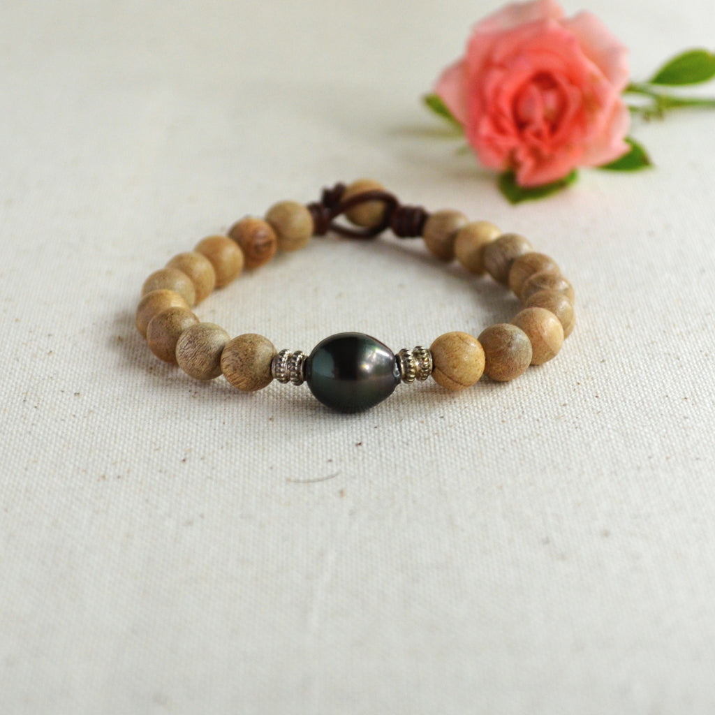 A beaded bracelet made of light brown sandalwood beads with a central dark black Tahitian pearl and small sterling silver spacer beads, displayed on a textured off-white fabric background with a pink rose in the background.