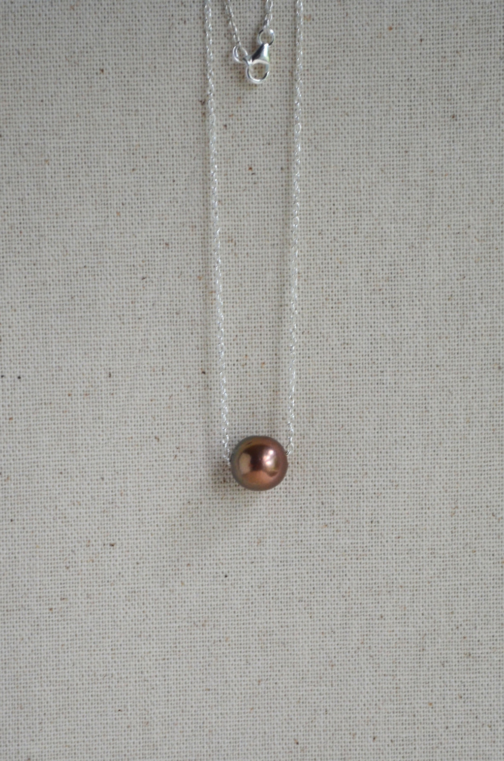 A delicate silver necklace with a single round, dark copper-brown pearl pendant, displayed against a textured beige linen background.