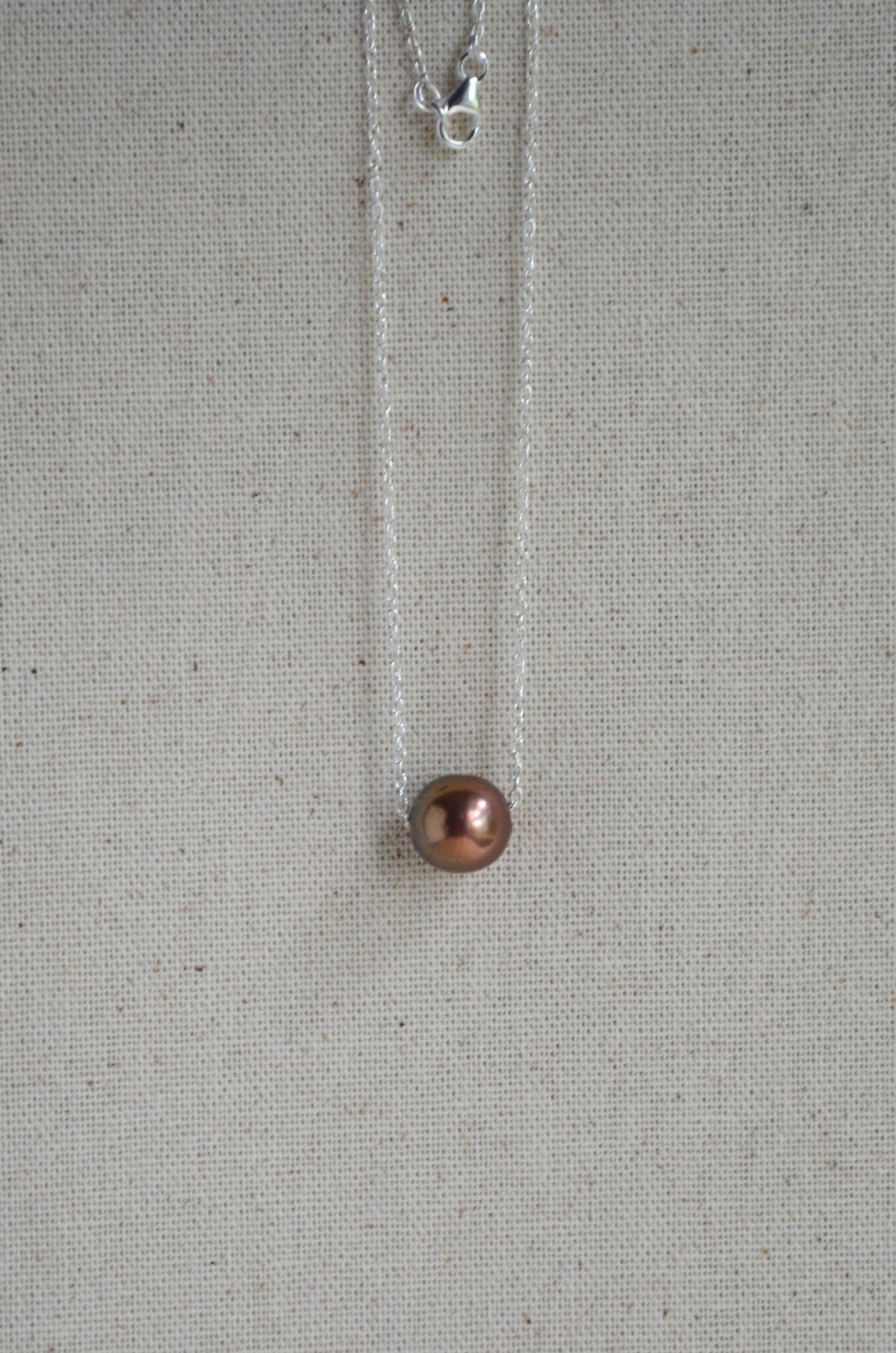 A delicate silver necklace with a single round, dark copper-brown pearl pendant, displayed against a textured beige linen background.
