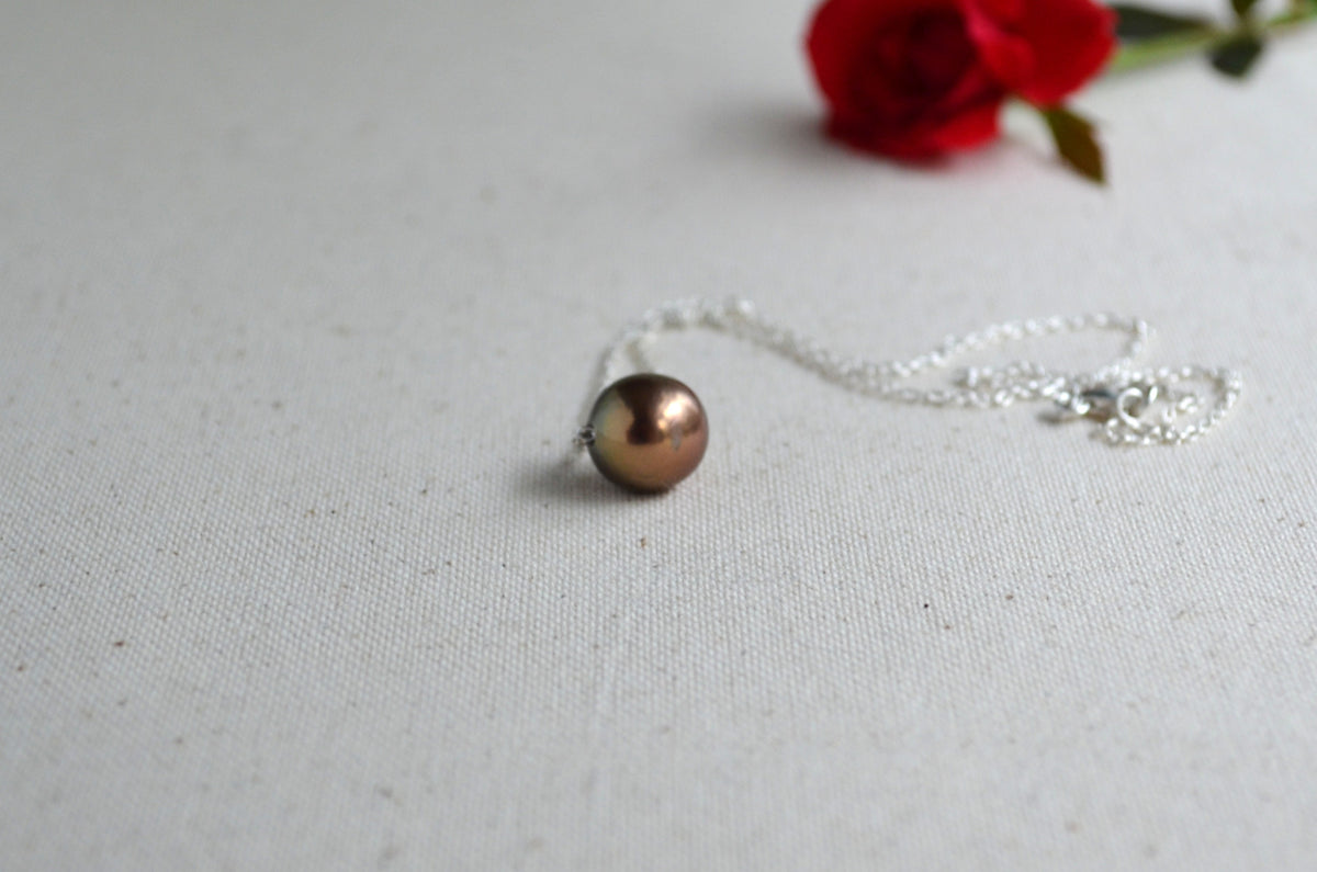 A silver necklace with a single round, metallic bronze-brown pearl pendant, displayed on a textured off-white fabric background with a blurred red rose in the upper right corner.
