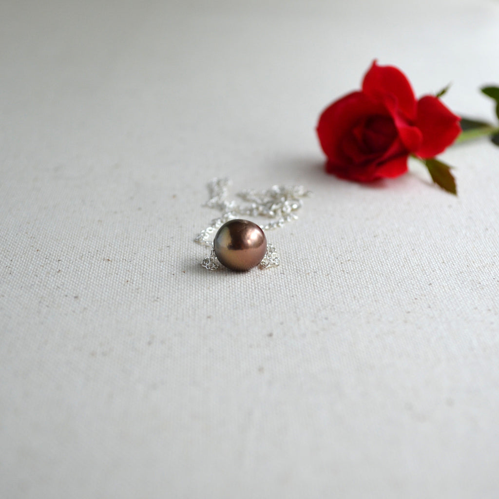 A sterling silver chain with a single oval brown Edison pearl, styled on a textured off-white fabric surface next to a red rose.
