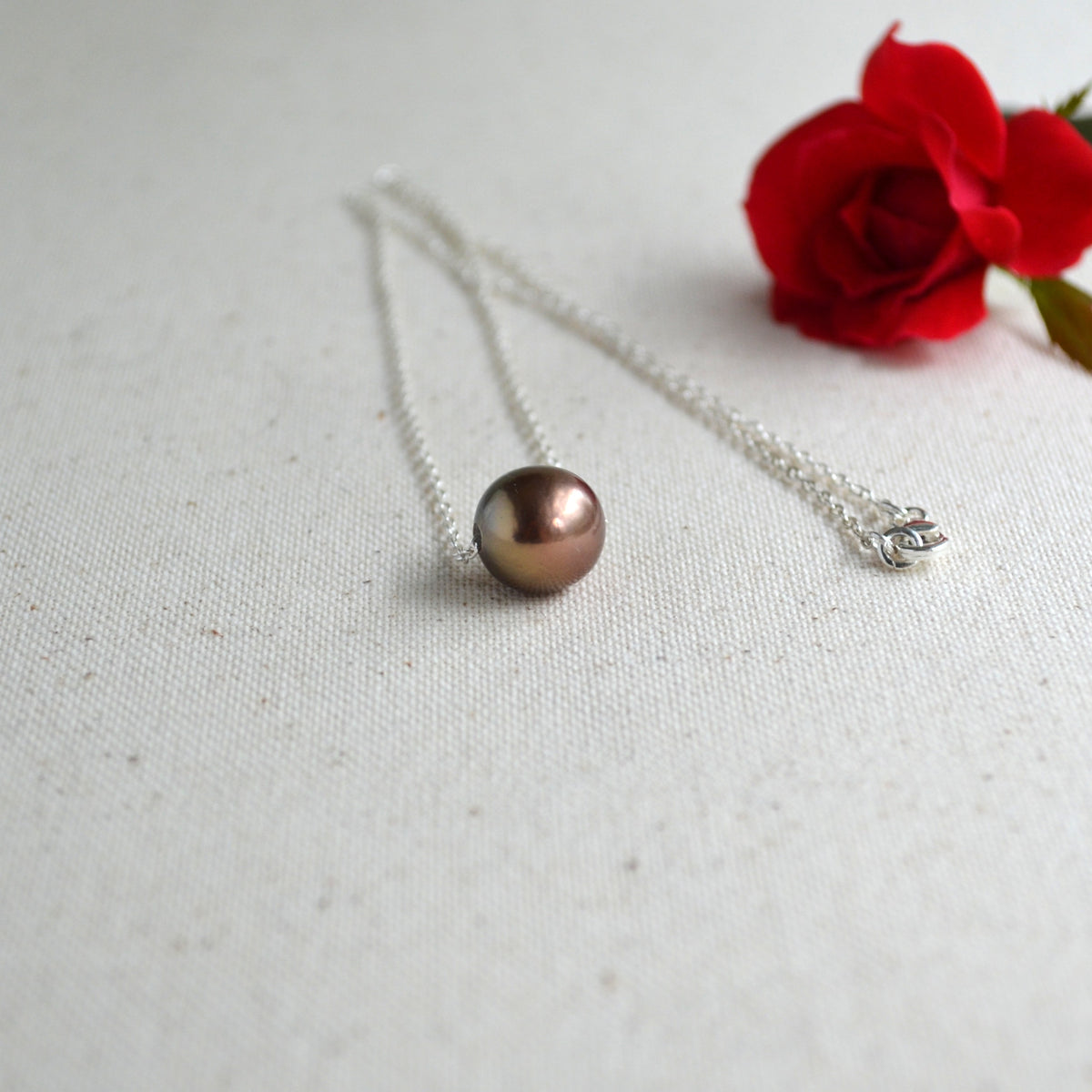 A silver chain necklace with a single round, dark metallic pearl pendant resting on a textured off-white fabric surface, with a red rose in the background.