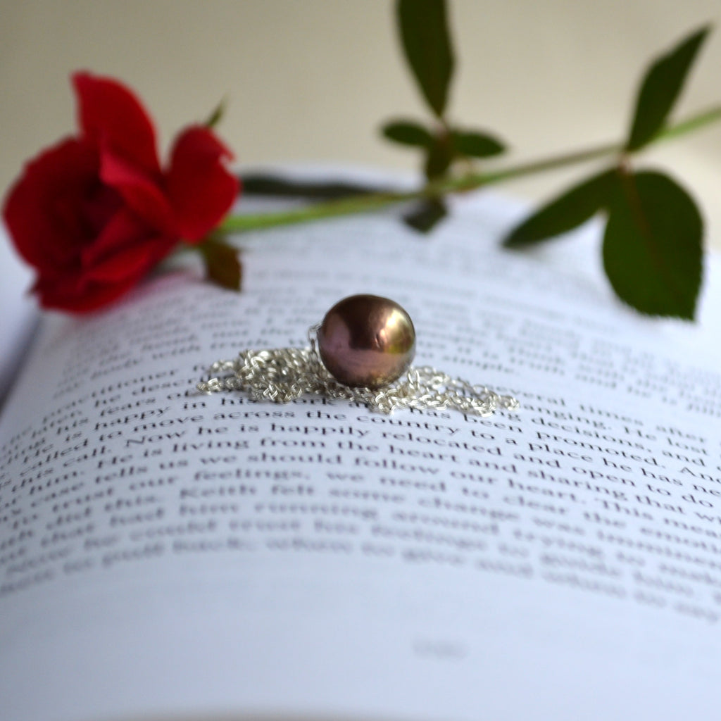 A silver chain necklace with a bronze-brown Edison pearl, resting on an open book with a red rose and green leaves in the background.