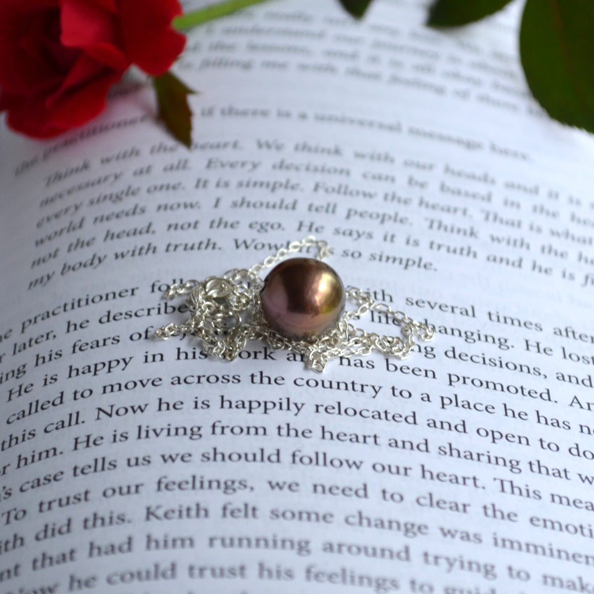 A sterling silver necklace with a round, chocolate Edison pearl, resting on an open book with blurred red flowers in the background.