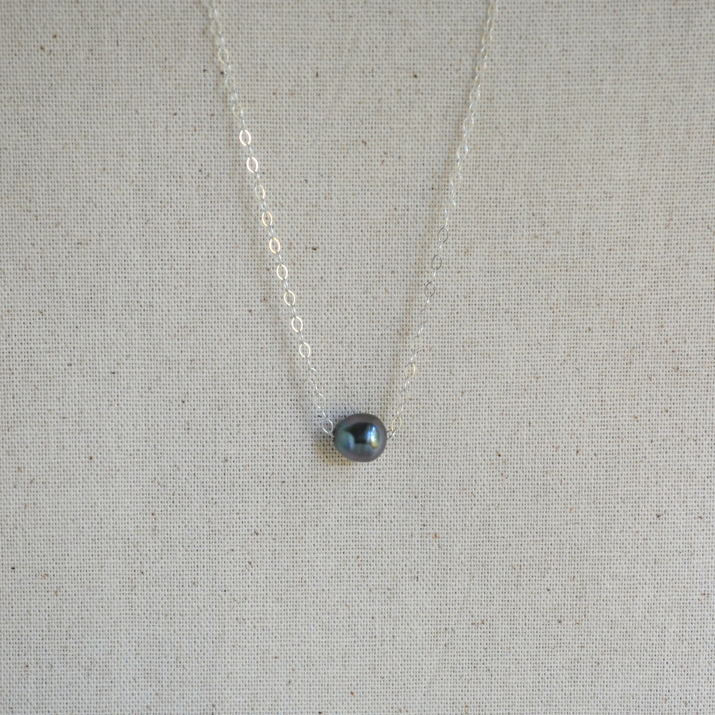 A delicate silver chain necklace with a small, teardrop-shaped dark gray iridescent Tahitian pearl pendant, displayed on a textured beige linen fabric background.