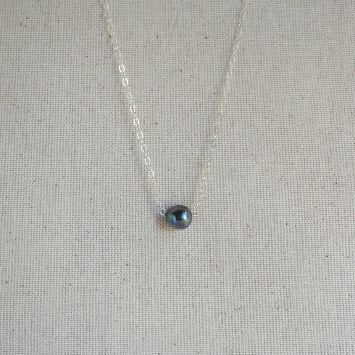 A delicate silver chain necklace with a small, teardrop-shaped dark gray iridescent Tahitian pearl pendant, displayed on a textured beige linen fabric background.