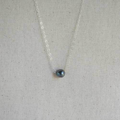 A delicate silver chain necklace with a small, teardrop-shaped dark gray iridescent Tahitian pearl pendant, displayed on a textured beige linen fabric background.