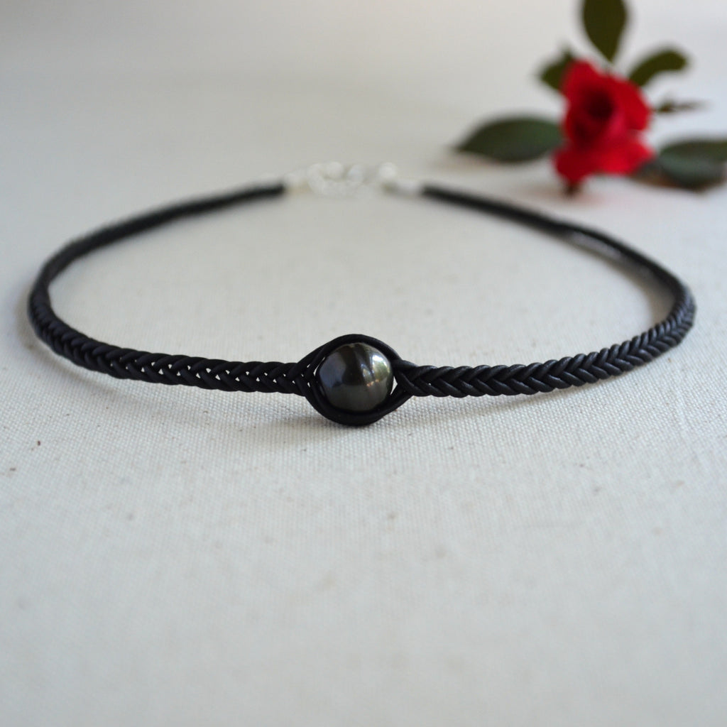 A black braided cord bracelet with a central round black bead or cabochon, photographed on a light neutral surface with a small red flower in the background.