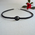 A black braided cord bracelet with a central round black bead or cabochon, photographed on a light neutral surface with a small red flower in the background.