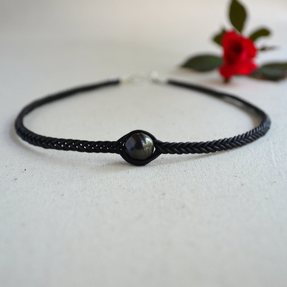 A black braided cord bracelet with a central dark gray pearl bead, displayed on a textured off-white fabric background with a blurred red rose in the background.