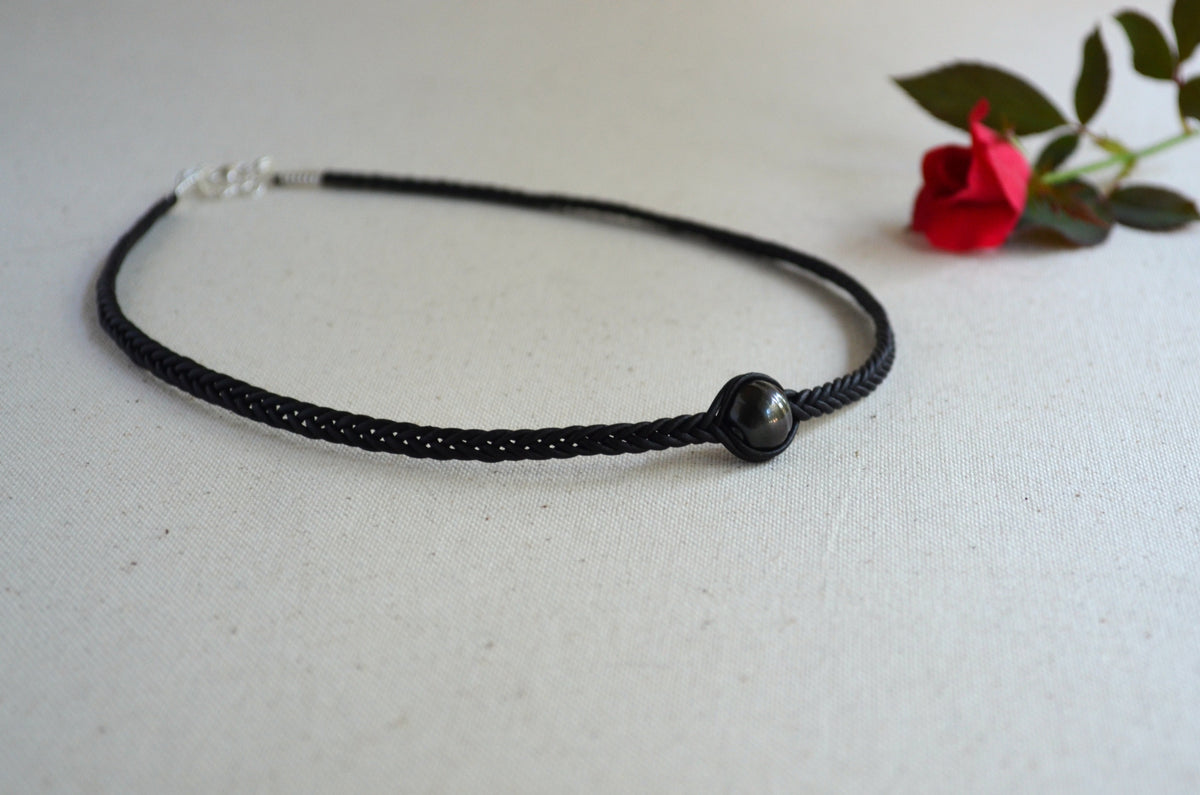 A black braided cord necklace with a single round black bead and a silver clasp, displayed on a light beige textured fabric surface with a red rose in the background.