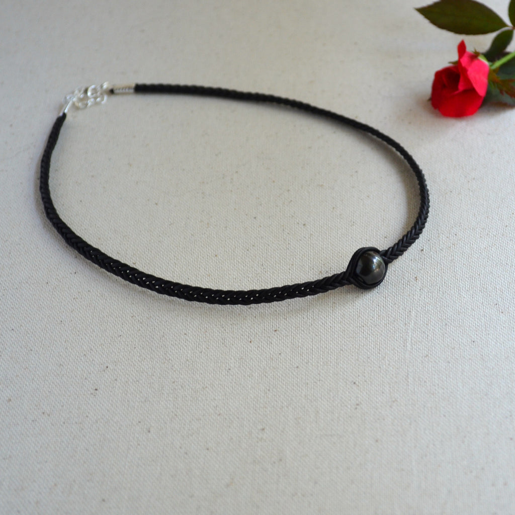 A black braided cord necklace with a single dark round bead and a silver clasp, displayed on a textured off-white fabric background with a small red rose in the corner.