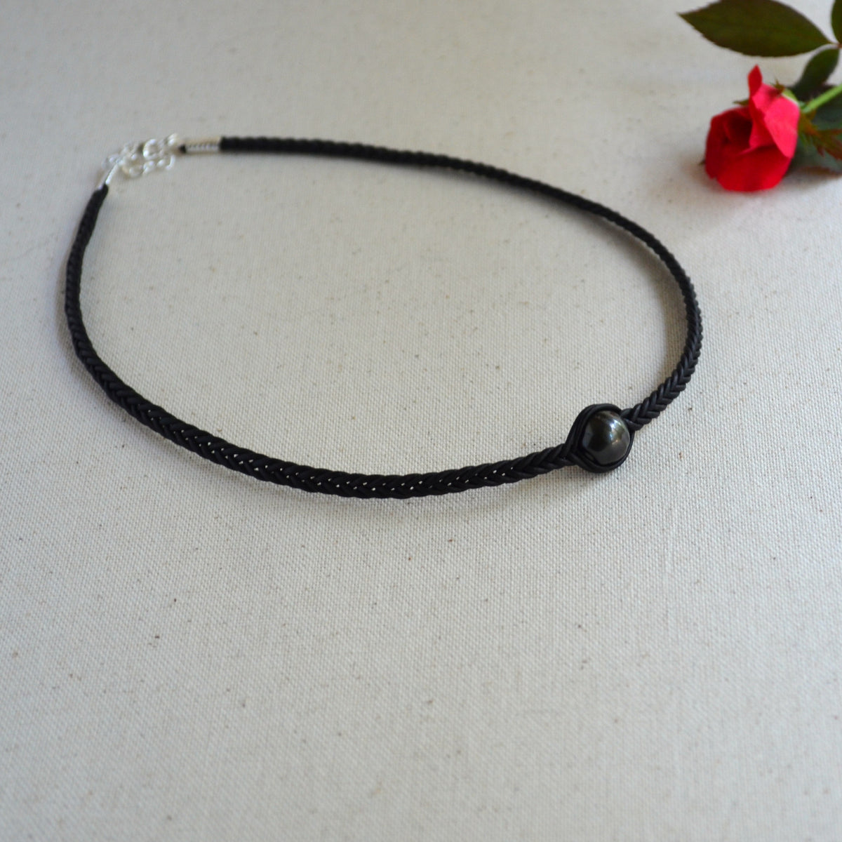 A black braided cord necklace with a single dark round bead and a silver clasp, displayed on a textured off-white fabric background with a small red rose in the corner.
