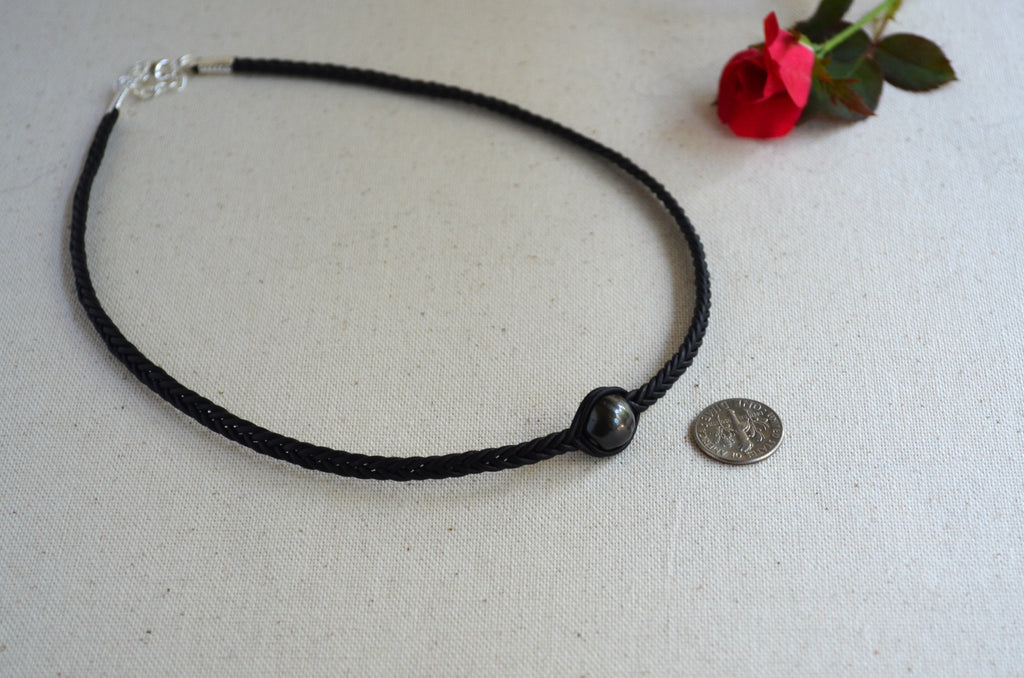 A black braided cord necklace with a single black bead and silver clasp, displayed on a light beige fabric surface next to a red rose and a coin for scale.