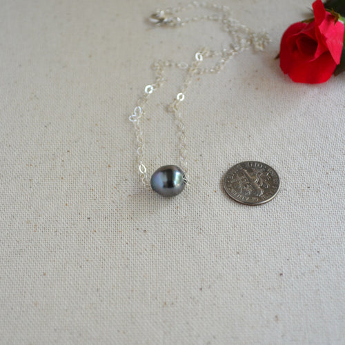 A silver chain necklace with a single dark gray pearl pendant, displayed on a textured beige fabric next to a coin for scale and a red rose in the corner.