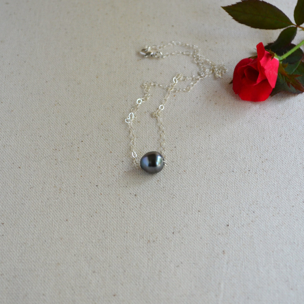 A delicate silver chain necklace with a single dark gray pearl pendant, styled on a textured beige fabric next to a red rose.