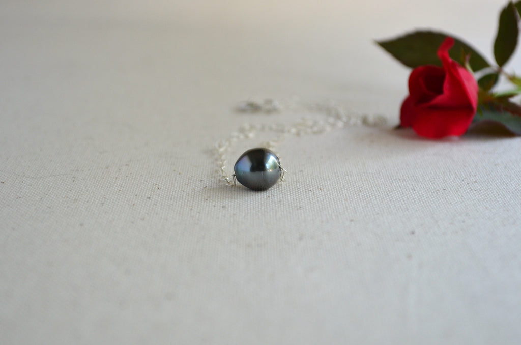 A silver ring with a round, dark gray-black pearl pendant, resting on a textured off-white fabric surface next to a red rose.