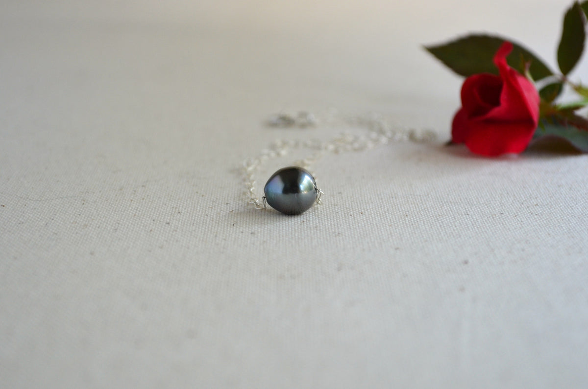 A silver ring with a round, dark gray-black pearl pendant, resting on a textured off-white fabric surface next to a red rose.