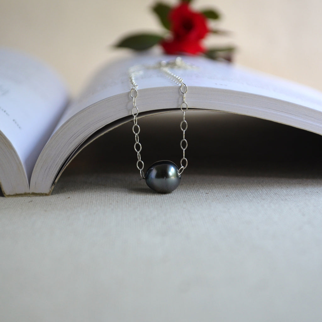 A silver chain necklace with a single dark gray pearl pendant, draped over the pages of an open book with a blurred red rose in the background.