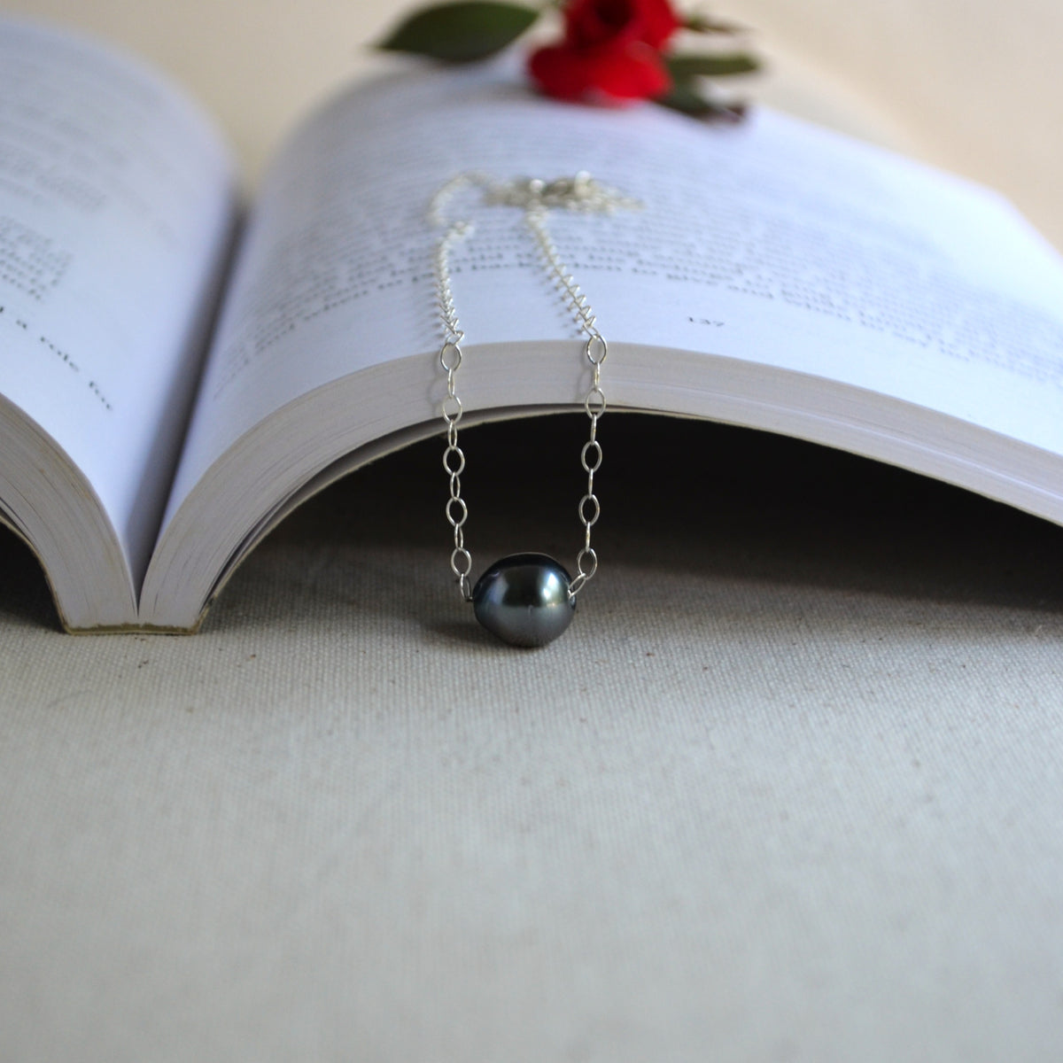A silver chain necklace with a single dark gray pearl pendant, draped over the pages of an open book with a small red flower accent.