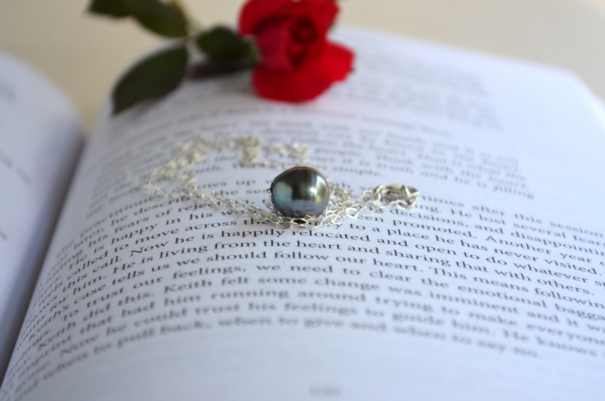 A delicate silver-toned necklace with a round dark gray pearl and a small gold heart accent, resting on an open book with a red rose in the background.