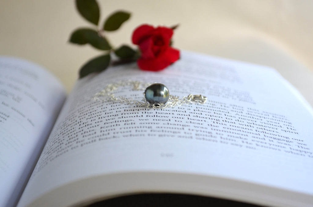 A silver-toned necklace with a dark, iridescent oval pendant rests on the pages of an open book, styled with a red rose and green leaves on a light beige background.