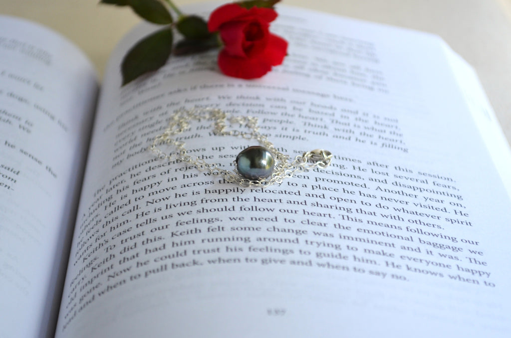 A silver-toned necklace with a dark gray teardrop-shaped gemstone and silver filigree, resting on the pages of an open book with a red rose tucked above it.