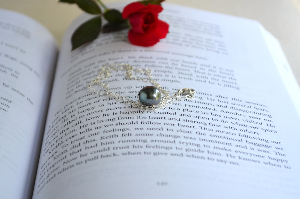 A silver-toned necklace with a dark gray teardrop-shaped gemstone and silver filigree, resting on the pages of an open book with a red rose tucked above it.
