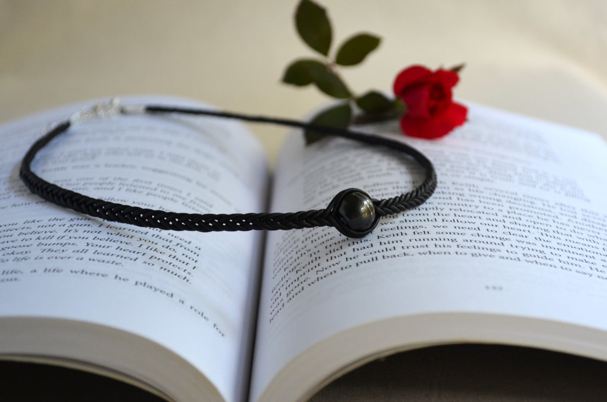 A black braided cord bracelet with a dark metallic bead centerpiece, resting on the pages of an open book with a red rose stem placed on top.