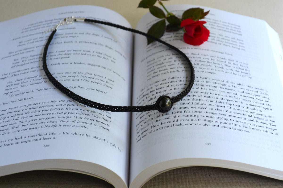 A black cord necklace with a dark gray round bead and silver-toned metal ends, laid across an open book with a red rose resting on the pages.