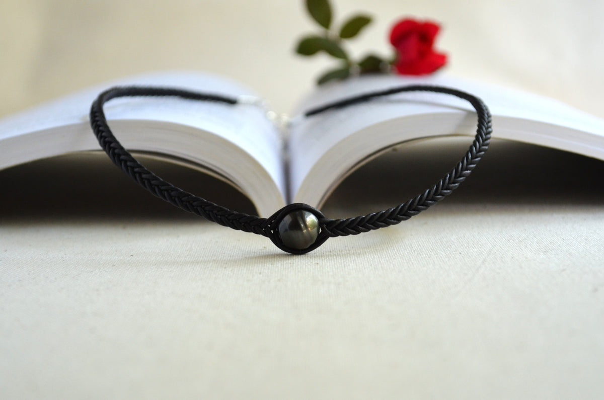 A black braided cord bracelet with a round dark metallic bead, displayed in front of an open book with a small red rose in the background.