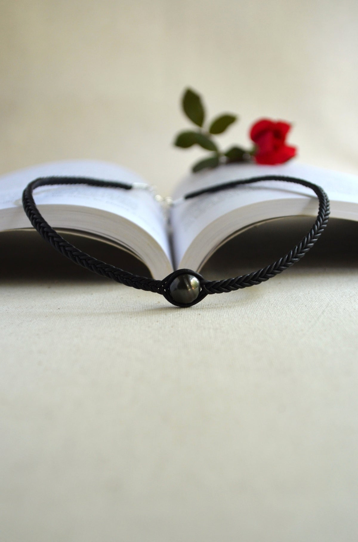 A black braided cord bracelet with a small dark metallic bead, displayed in front of an open book with a red rose on a light beige background.