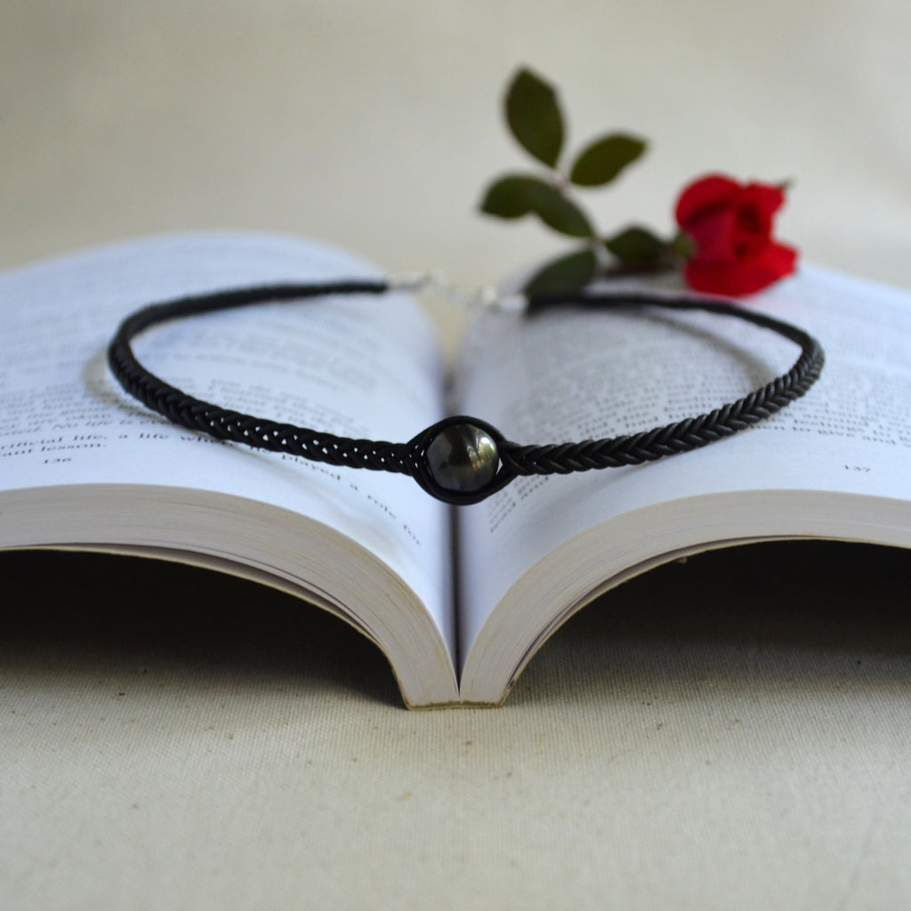 A black braided cord bracelet with a central glossy black bead, displayed on the pages of an open book with a small red rose resting above it.