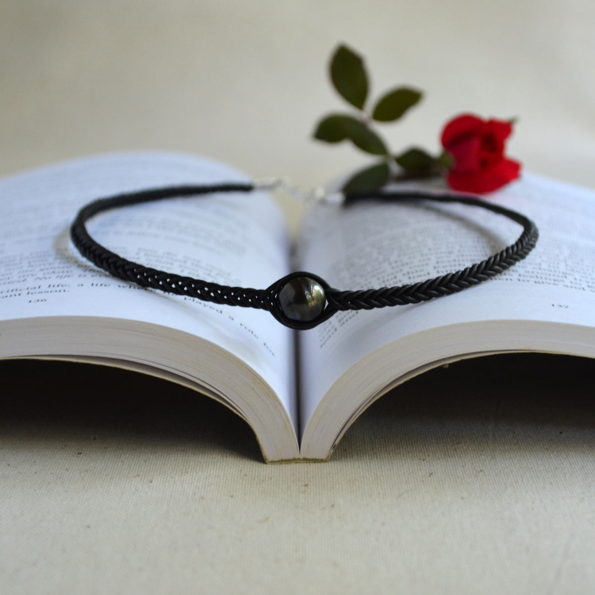 A black braided cord bracelet with a central glossy black bead, displayed on the pages of an open book with a small red rose resting above it.