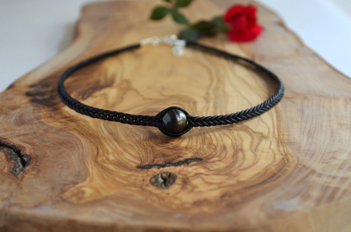 A black braided cord bracelet with a central polished dark stone bead, displayed on a rustic wooden board with a small red rose in the background.