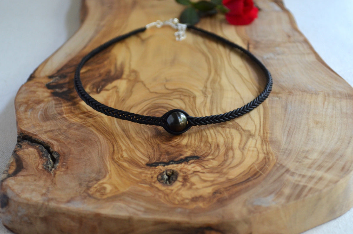 A black braided cord necklace with a single round black bead centerpiece, displayed on a rustic wood slice with a small red flower in the corner.